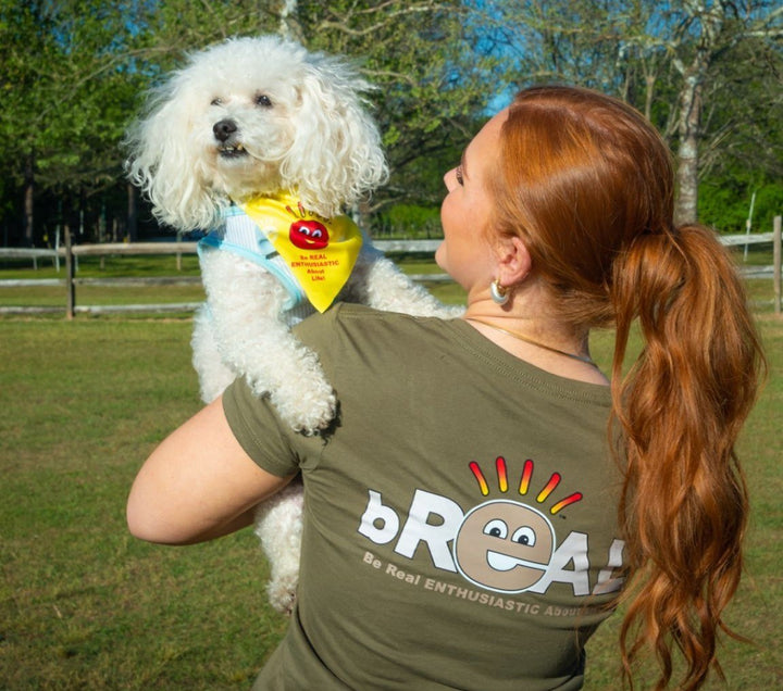 A woman in a bREAL tee holding up her fluffy white dog wearing a yellow bREAL bandana