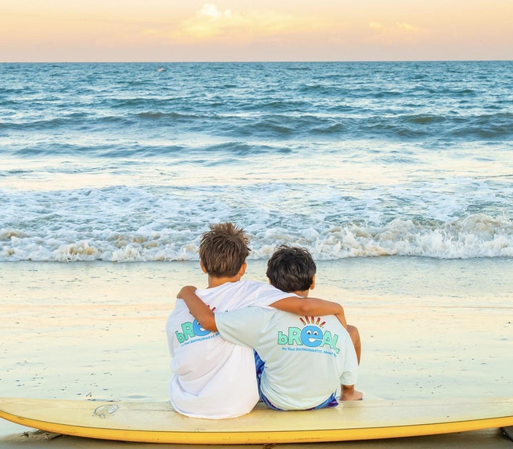 Two kids sitting arm in arm on a surfboard at the beach, both wearing bREAL tees