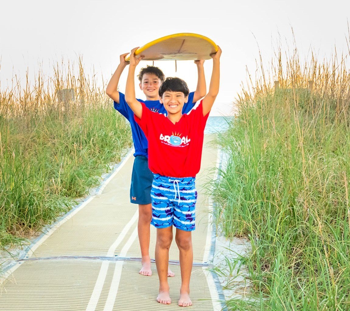 Two brothers holding a surfboard over their heads on a beach boardwalk, one in a red bREAL tee